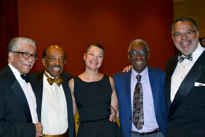four men and one woman standing side by side posing for a photo with their arms around each other, they are all dressed up