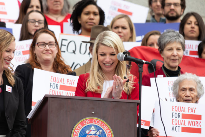 A large group of women holding up equal rights signs behind a female legislator speaking at a podium