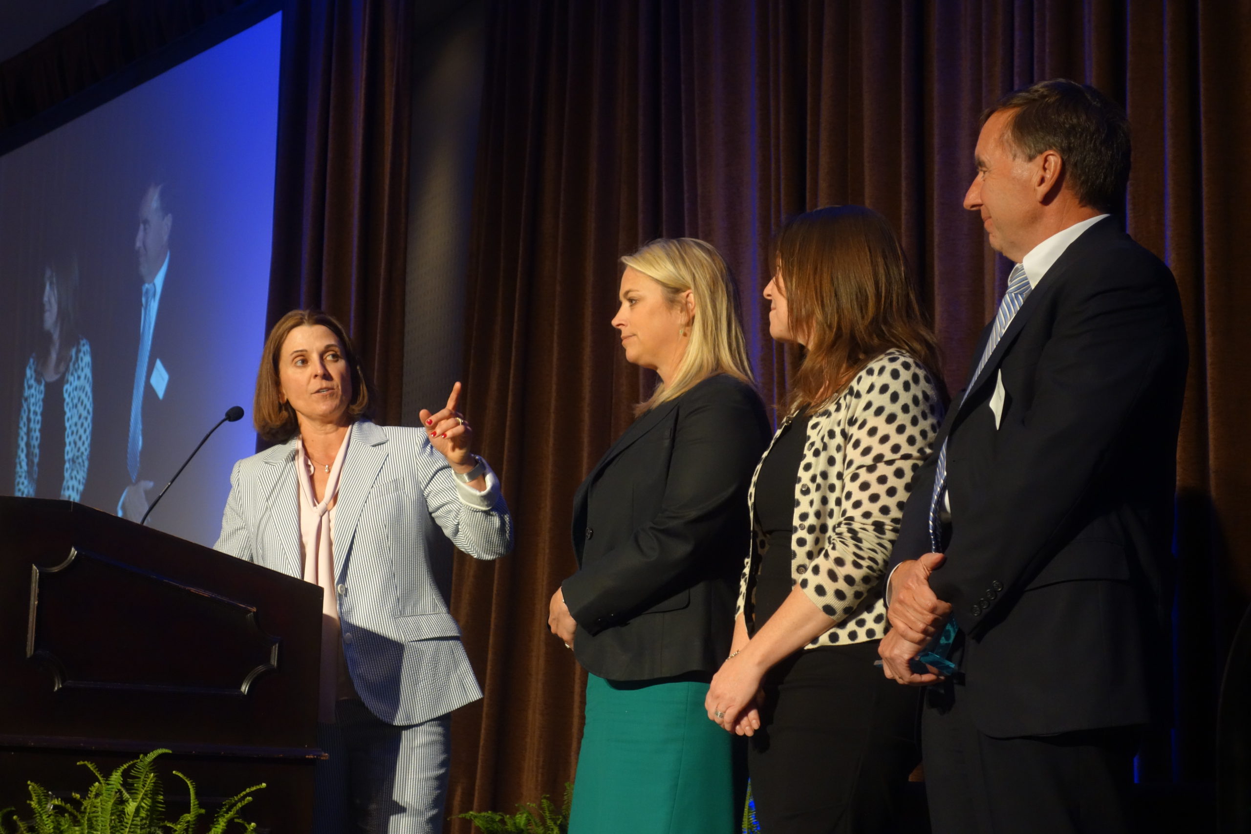 A female speaker on a stage pointing to three individuals being recognized for an accomplishment