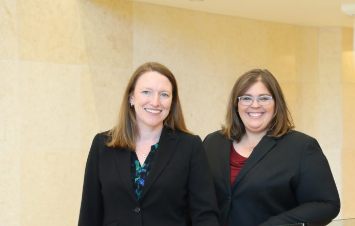 Two standing shoulder to shoulder in black suits smiling in front of a beige wall