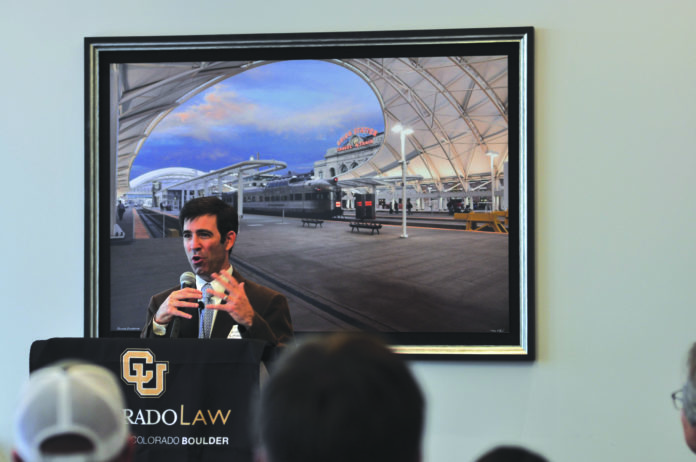 A man in a suit speaking at a crowdfunding event in front of a large photo of a train station