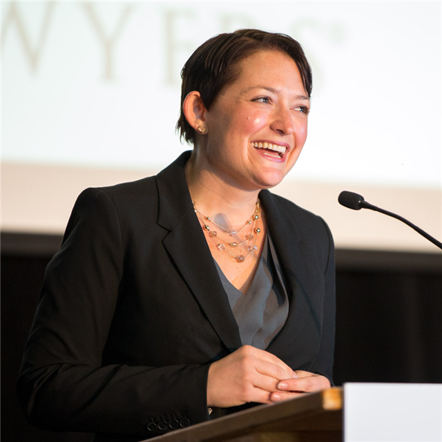 A woman with a short hair a black blazer and a big smile on her face speaking at an Educating Tomorrow’s Lawyers conference