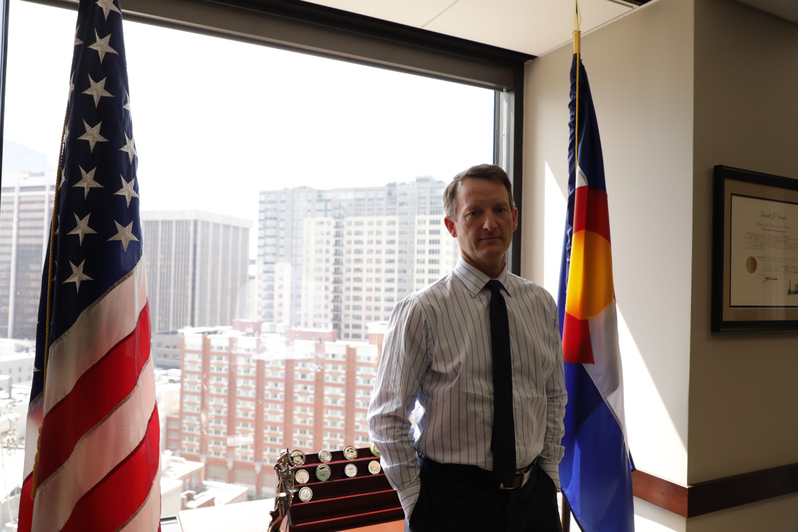 A man in a white button up and black tie in front of a big window with two American flags on either side of him