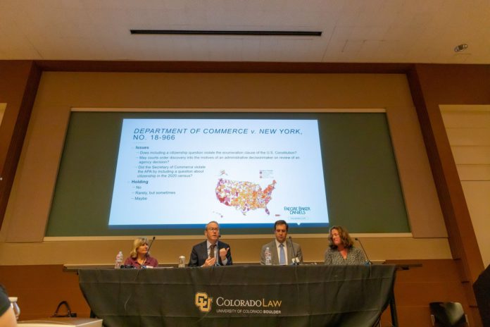 Four people sitting at a table on a stage in front of a large projector screen Four people sitting at a table on a stage in front of a large projector screenFour people sitting at a table on a stage in front of a large projector screen