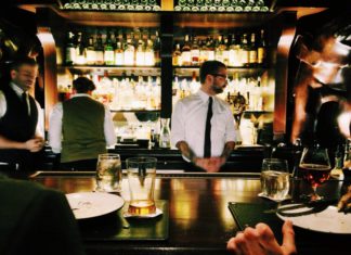Overtime Ceiling Set to Rise in January A Bartender in a white button up and black tie stands behind a bar waiting for his next order