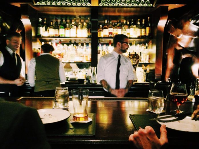 A Bartender in a white button up and black tie stands behind a bar waiting for his next order