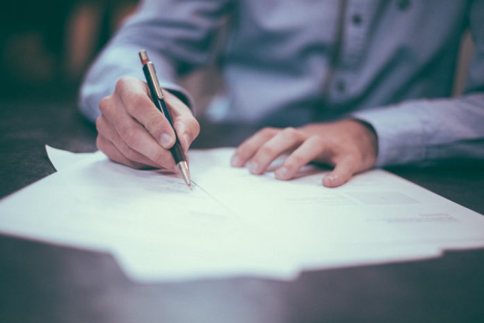 A man in a dress shirt writes on forms scattered on a desktop.