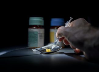 AG’s Office Reveals All Municipalities Eligible for Opioid Settlement Funds A hand holding a needle hovers over drugs resting in a spoon on a table with pill bottles in view.