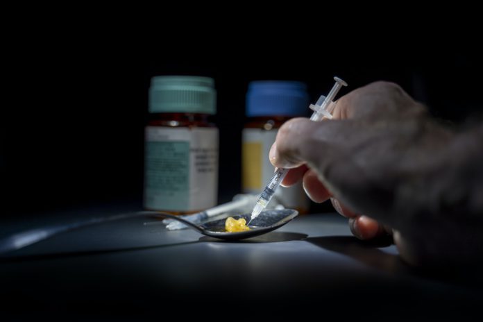 A hand holding a needle hovers over drugs resting in a spoon on a table with pill bottles in view.