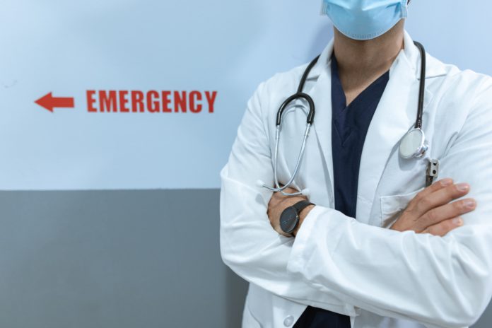 A doctor stands with arms crossed near a sign for the emergency room.