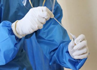 AG Phil Weiser Orders 11 COVID Testing Sites to Shut Down Person in personal protective equipment places swab in test tube.