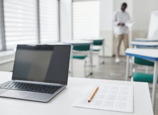 Colorado Bar Exam Returns In-Person With COVID Precautions An open laptop, a blank multiple choice sheet and a pencil sit on a desk in an empty classroom with a man standing in the background.