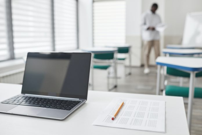 An open laptop, a blank multiple choice sheet and a pencil sit on a desk in an empty classroom with a man standing in the background.