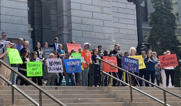 A group of people hold signs behind a man speaking at a podium on top of stone steps