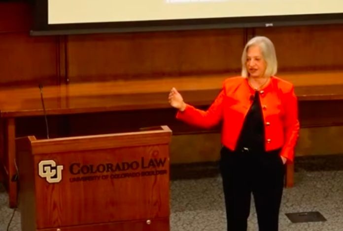 A woman with short, gray hair in an orange blazer speaking next to a podium with the University of Colorado Law School printed on it.