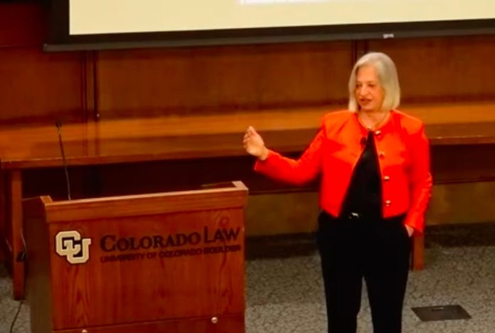 Lecture A woman with short, gray hair in an orange blazer speaking next to a podium with the University of Colorado Law School printed on it.