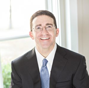 Man with glasses in black suit with blue tie smiling 