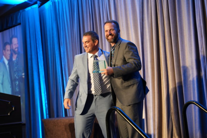 CM100251 A man in a light gray suit with his arm on a man in a dark gray suit who is smiling at the camera and holding a glass award on a stage.