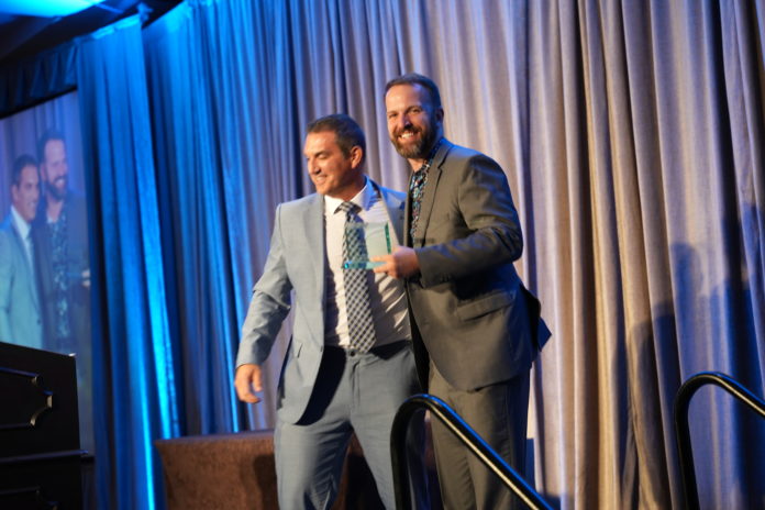 A man in a light gray suit with his arm on a man in a dark gray suit who is smiling at the camera and holding a glass award on a stage.