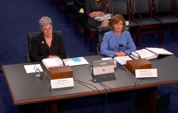 May12Hearing A woman with short white hair in a black suit sits to the left of a woman with short brown hair in a light blue suit in front of a large table.