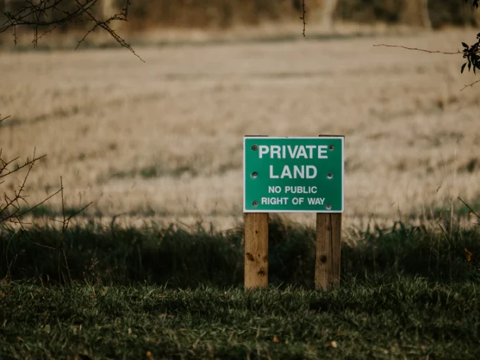 A green sign that reads “Private Land No Right of Way” with bullet holes in it in front of an open field.