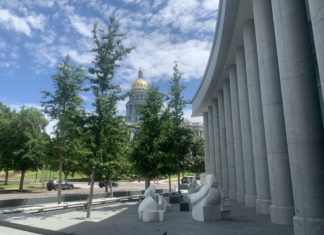 Possible Expansion for Colorado Program Bridging Criminal Justice and Mental Health A building with a golden dome on top of a detailed rotunda is visible behind trees and cars with columns from another building in the foreground and a white stone statue of three figures looking at another figure.