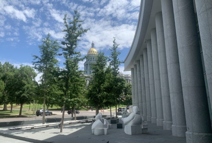 A building with a golden dome on top of a detailed rotunda is visible behind trees and cars with columns from another building in the foreground and a white stone statue of three figures looking at another figure.