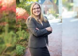 Brittany Kauffman Named IAALS CEO A blonde woman in glasses wearing a gray skirt suit smiles and crosses her arms in front of red brick buildings.