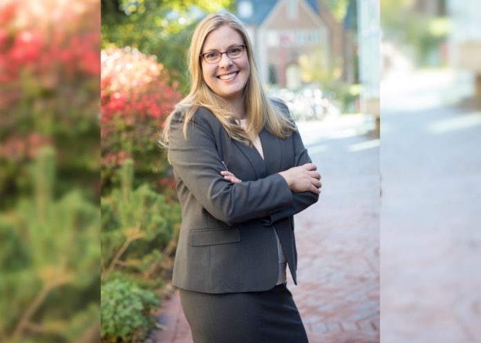 A blonde woman in glasses wearing a gray skirt suit smiles and crosses her arms in front of red brick buildings.