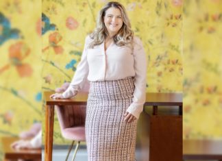 Paloma Law Hopes to Improve Access to Justice on the Ground A blonde woman in a pencil skirt and button up shirt stands with one hand on a wooden desk and smiles at the camera. The background has yellow floral wall paper and a pink velvet chair.
