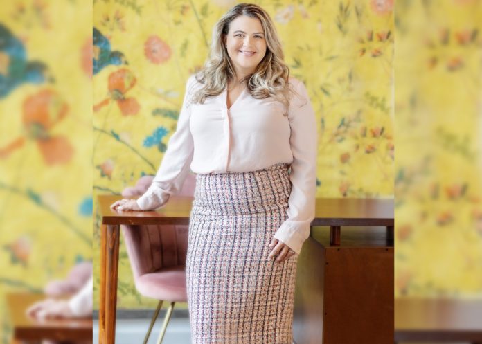 Erica White - Paloma Law A blonde woman in a pencil skirt and button up shirt stands with one hand on a wooden desk and smiles at the camera. The background has yellow floral wall paper and a pink velvet chair.