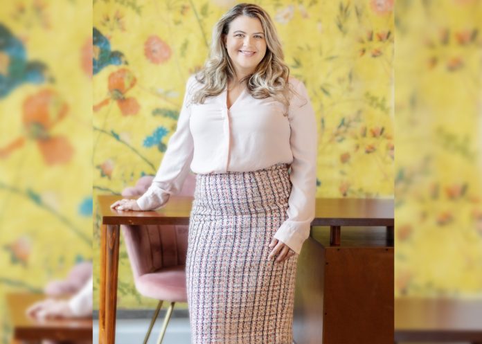 A blonde woman in a pencil skirt and button up shirt stands with one hand on a wooden desk and smiles at the camera. The background has yellow floral wall paper and a pink velvet chair.