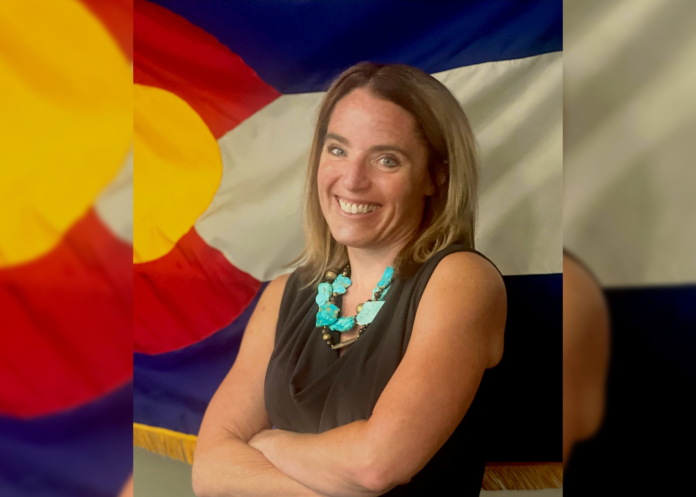 A woman stands in front of a Colorado state flag smiling wearing a brown top.