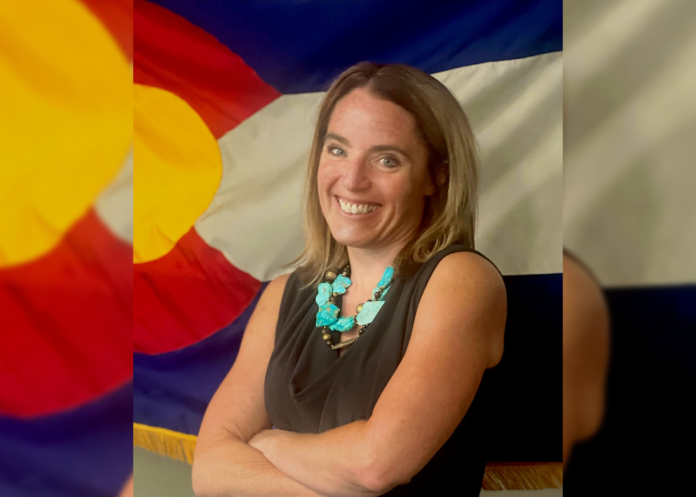 Anne Kelly A woman stands in front of a Colorado state flag smiling wearing a brown top.