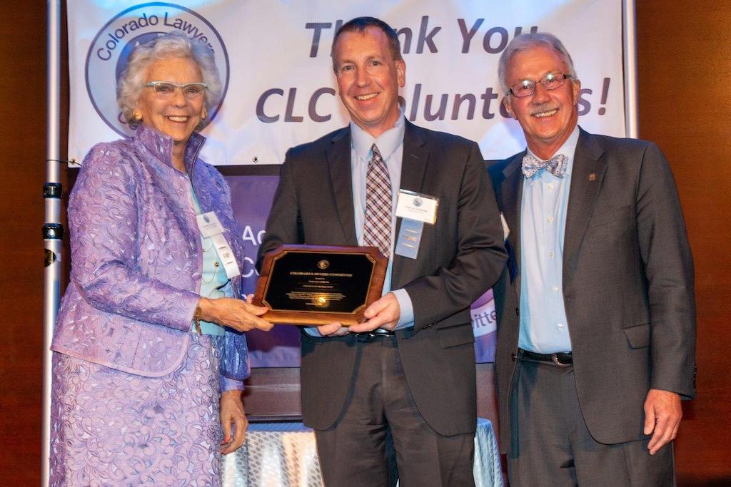 Three people, an older woman in a purple suit, a middle aged man in a suit and an older man in a suit, hold a plaque and smile in front of a banner that reads “Thank You CLC Volunteers.” 