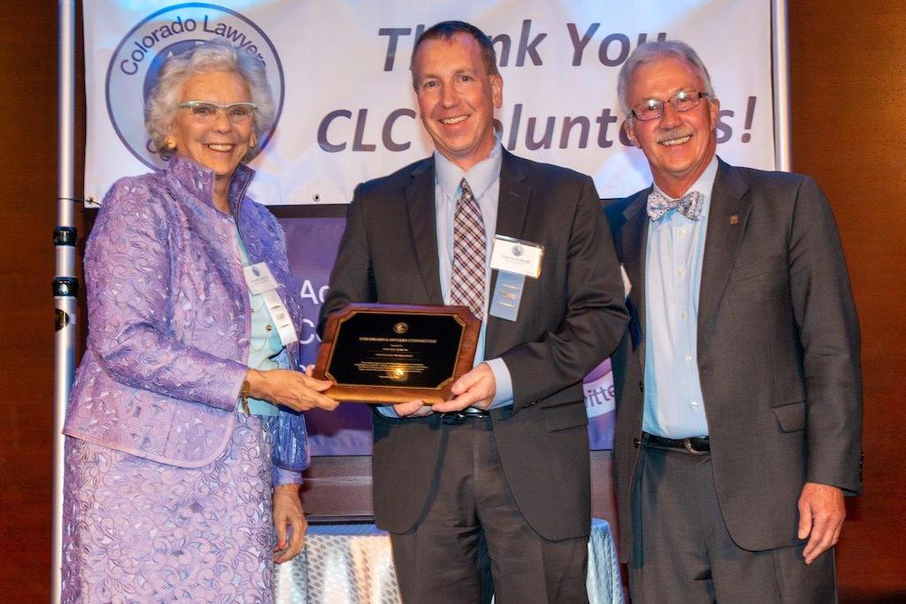 Three people, an older woman in a purple suit, a middle aged man in a suit and an older man in a suit, hold a plaque and smile in front of a banner that reads “Thank You CLC Volunteers.”