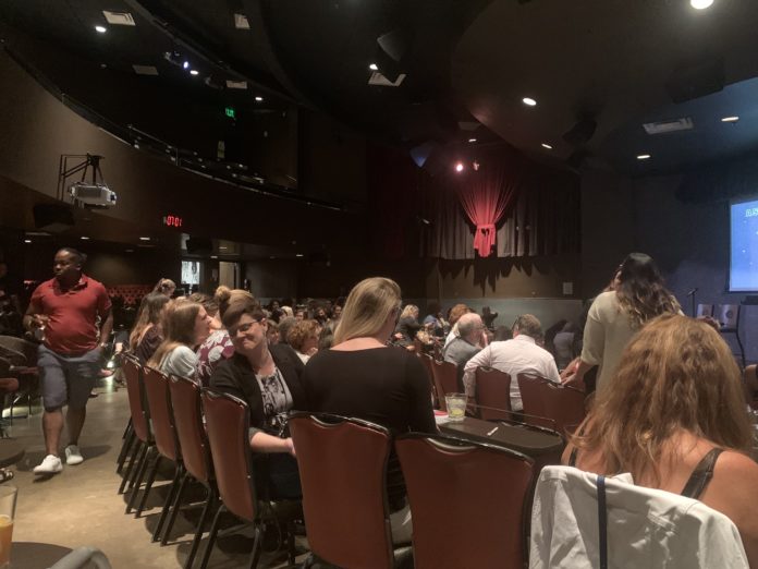 An empty stage surrounded by a crowd of people sitting in chairs in front of small tables.