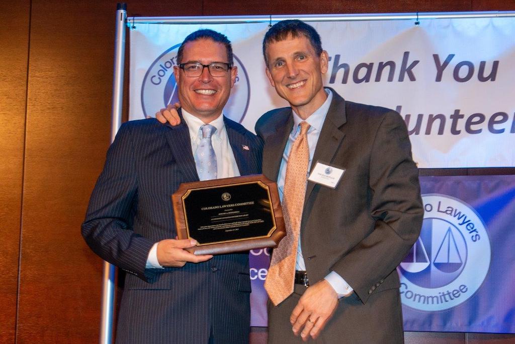 Two middle aged men in suits smile. The one on the left in a dark pinstripe suit and glasses holds a plaque. The one on the right in a brown suit has his hand on the other’s shoulder. 