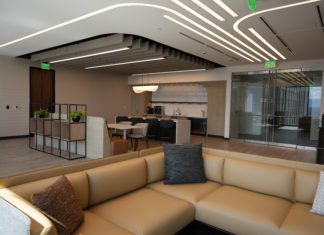 Lathrop GPM’s New Denver Office is Embracing the Future of Work A large open office space. The foreground has a tan, leather sectional couch with throw pillows. In the back, a small kitchen with stainless steel appliances and a sleek white island with leather chairs. Curved lighting is embedded in the ceiling.