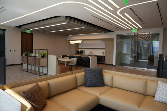 A large open office space. The foreground has a tan, leather sectional couch with throw pillows. In the back, a small kitchen with stainless steel appliances and a sleek white island with leather chairs. Curved lighting is embedded in the ceiling.