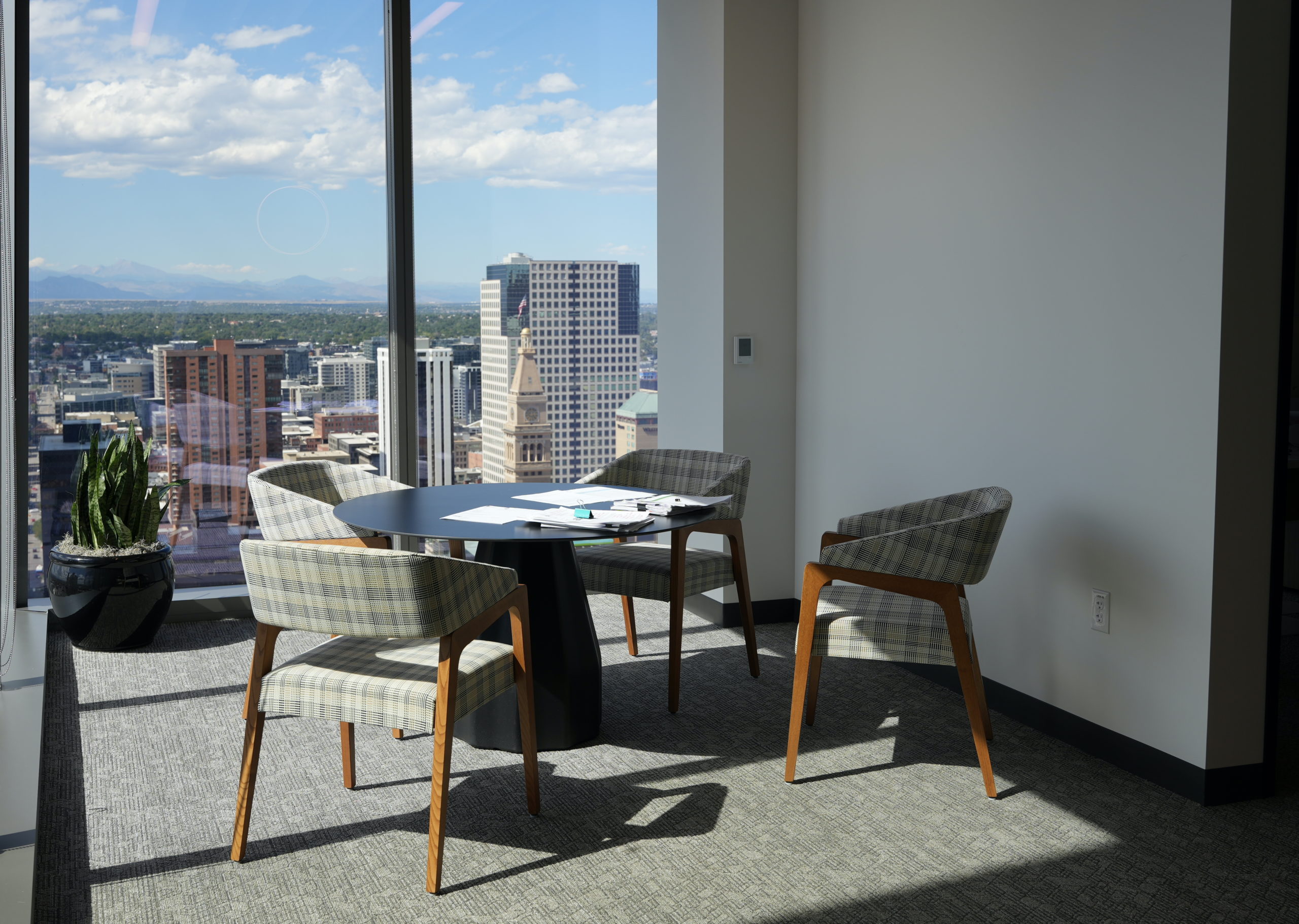 A round table with four cloth chairs around it and papers on top is in an office alcove. Two sides of the alcove are floor length glass windows. Outside, is a view of downtown Denver with mountains in the background.