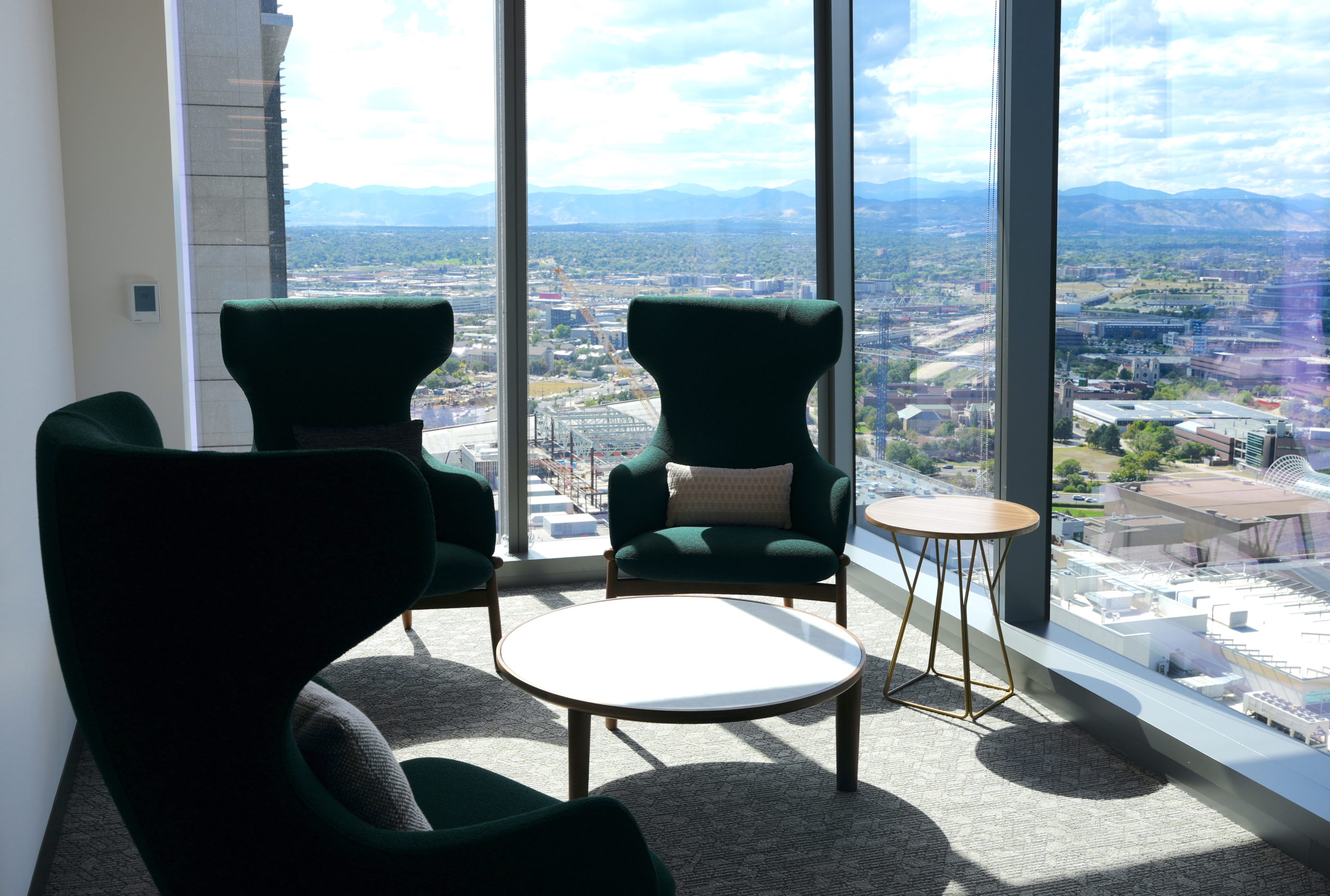 Three high-backed dark green cloth chairs with throw pillows face each other in an office alcove. An empty coffee table is in the middle. Two sides of the alcove are floor length glass windows. Outside, is a view of West Denver with mountains in the background.