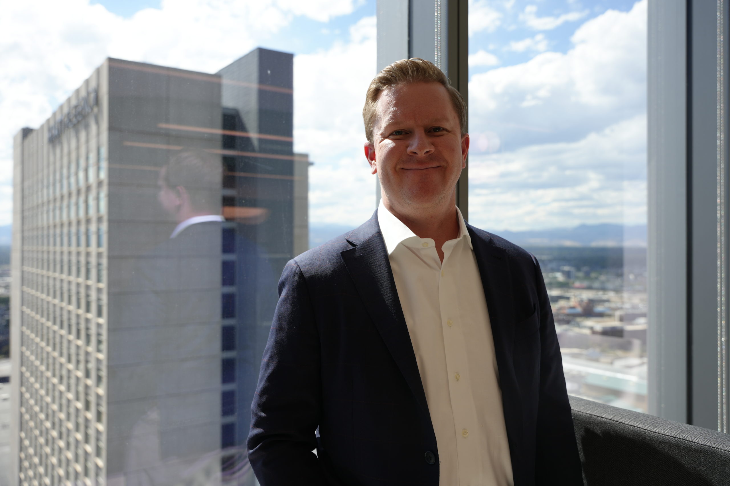 A middle- aged man with red hair wearing a gray suit and white button up shirt smiles slightly in front of large glass windows with skyscrapers and distant mountains in the back. 