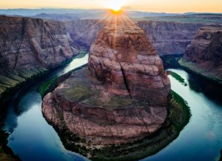 Where it Stands Now: 100th Anniversary of Colorado River Compact The Colorado River bends around an orange and gray canyon with green vegetation near the shoreline. The sun can be seen in the background overlooking the mountains.