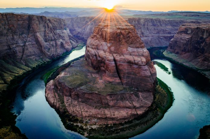 The Colorado River bends around an orange and gray canyon with green vegetation near the shoreline. The sun can be seen in the background overlooking the mountains.