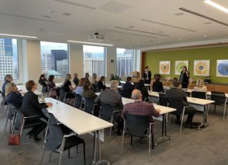 Colorado Judicial Institute Continues to Spread Legal Knowledge Two people stand in front of a group of people who are sitting at four different rows of tables. A city skyline can be seen in the background.