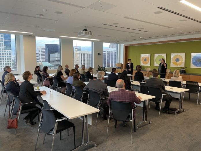 Two people stand in front of a group of people who are sitting at four different rows of tables. A city skyline can be seen in the background.