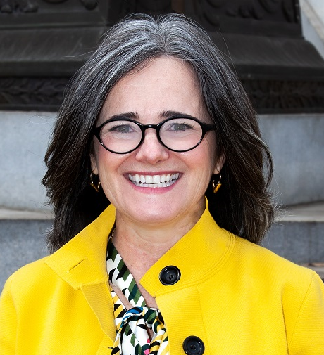 A woman in a yellow coat stands in front of some concrete steps. She wears glasses.