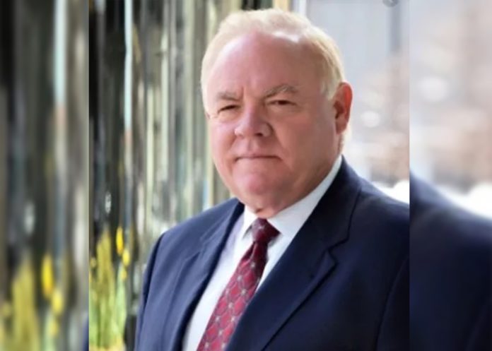 An older man in a suit looks at the camera with a stern expression.