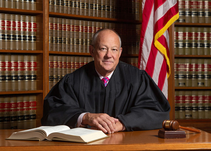 A man sits at a desk wearing a black judge’s robe with legal books behind him and an American flag.